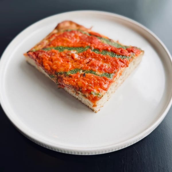 A thick slice of focaccia pizza with red sauce and green pesto stripes on a white plate.