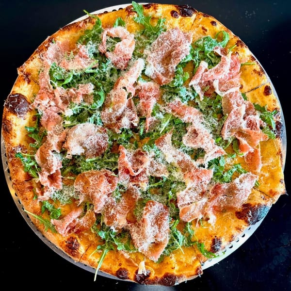 A whole pizza topped with prosciutto, fresh arugula, and grated Parmesan cheese on a dark surface.