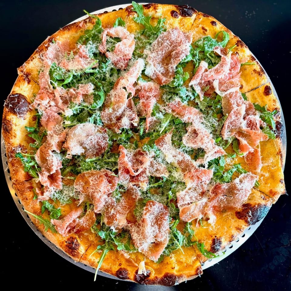 A whole pizza topped with prosciutto, fresh arugula, and grated Parmesan cheese on a dark surface.